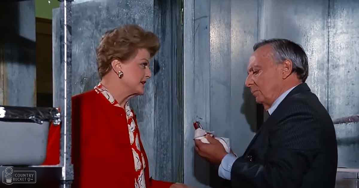A middle-aged female wearing a red cardigan and floral blouse with gold earrings, alongside an older adult male in a dark suit and light blue shirt holding a small white object indoors against metallic walls.