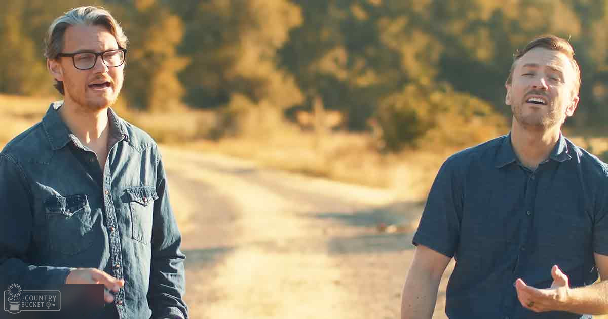 Two young adult males wearing casual shirts outdoors on a dirt road with late summer or early fall foliage and warm lighting.