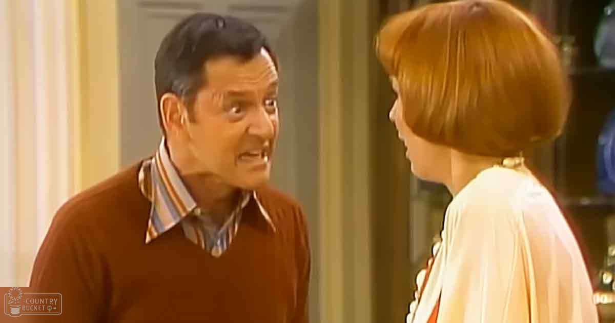 Middle-aged man wearing a brown, striped collared shirt under a relaxed fit long-sleeved sweater with cream/off-white, textured blouse or light jacket in an indoor home or office setting.