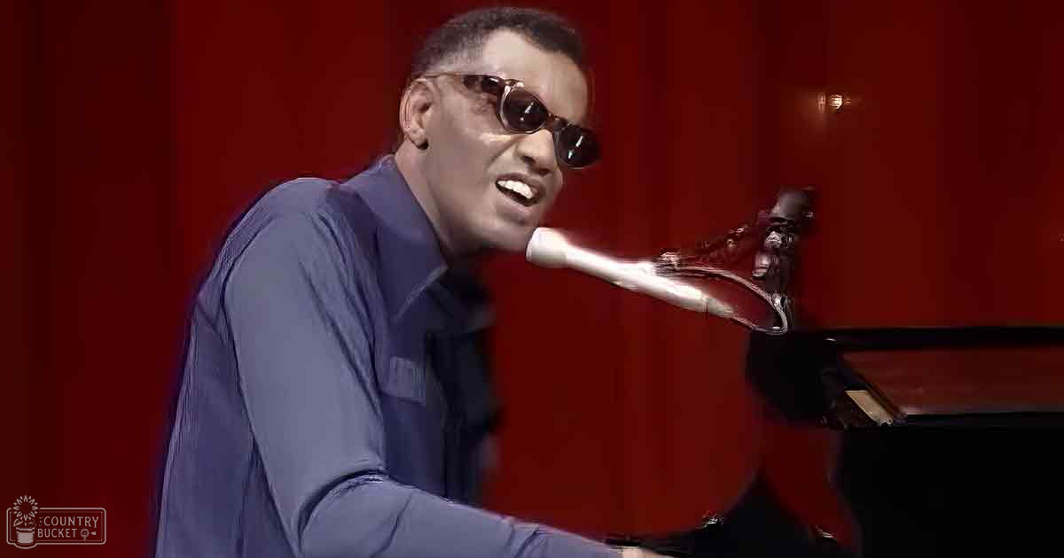 An adult male singer playing piano in a dimly lit indoor setting with red curtains, wearing a dark blue or navy button-down shirt and classic-style sunglasses.