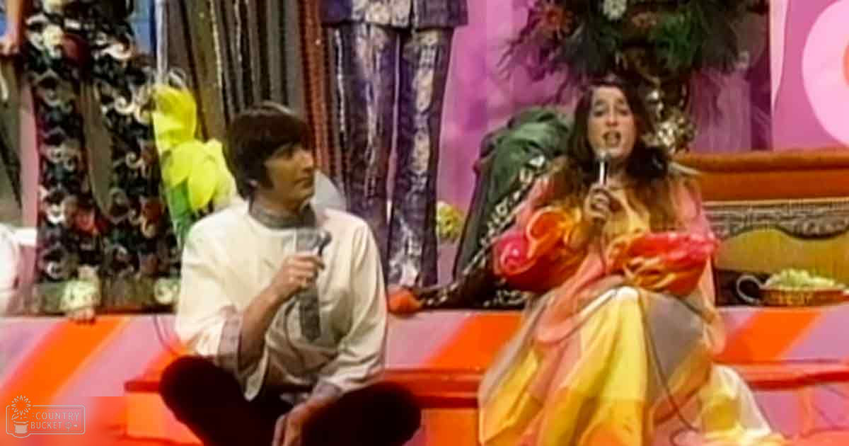 A male performer dressed in a white tunic and dark tie stands to the left holding a microphone; an adult female performer in a vibrant orange and yellow feathered dress stands to the right, also holding a microphone. The background suggests a theatrical stage with colorful curtains and floral arrangements.