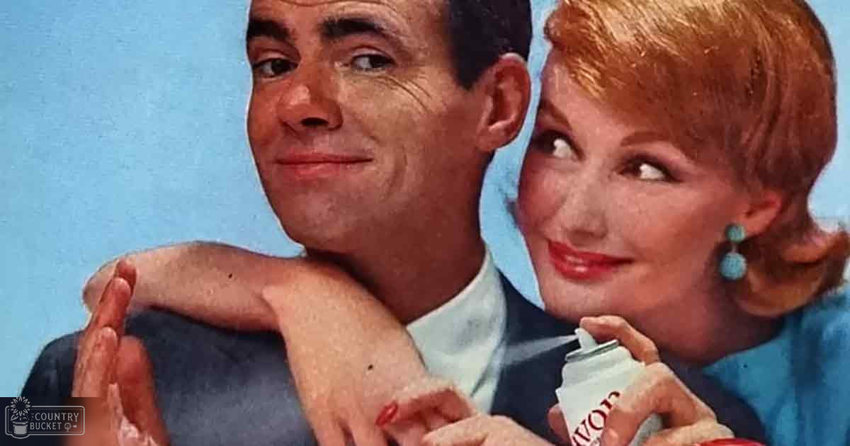 An adult male singer wearing a dark suit jacket with a light blue shirt, accompanied by an adult female in a blue dress or blouse with turquoise earrings and red nail polish, holding a small white Avon bottle against a plain light blue background.