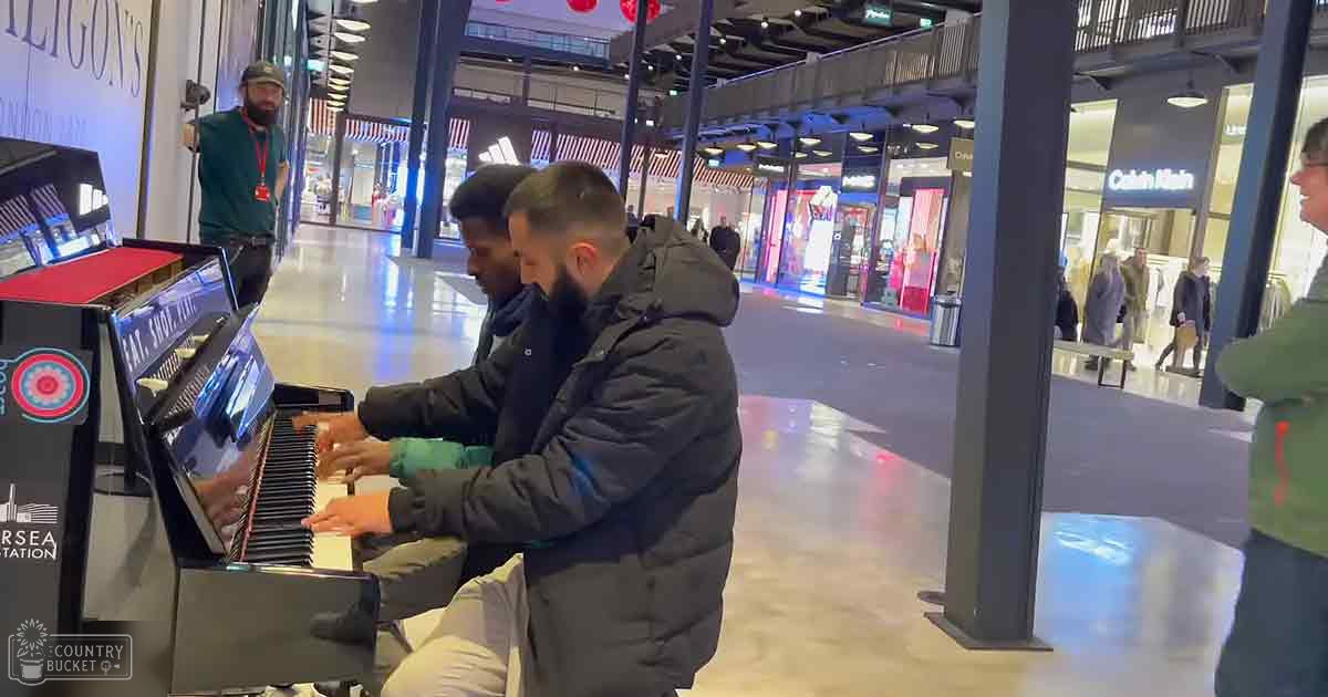 Two adult males play a digital piano in a shopping mall with visible Calvin Klein signage and other stores. A person in green attire records or observes nearby.