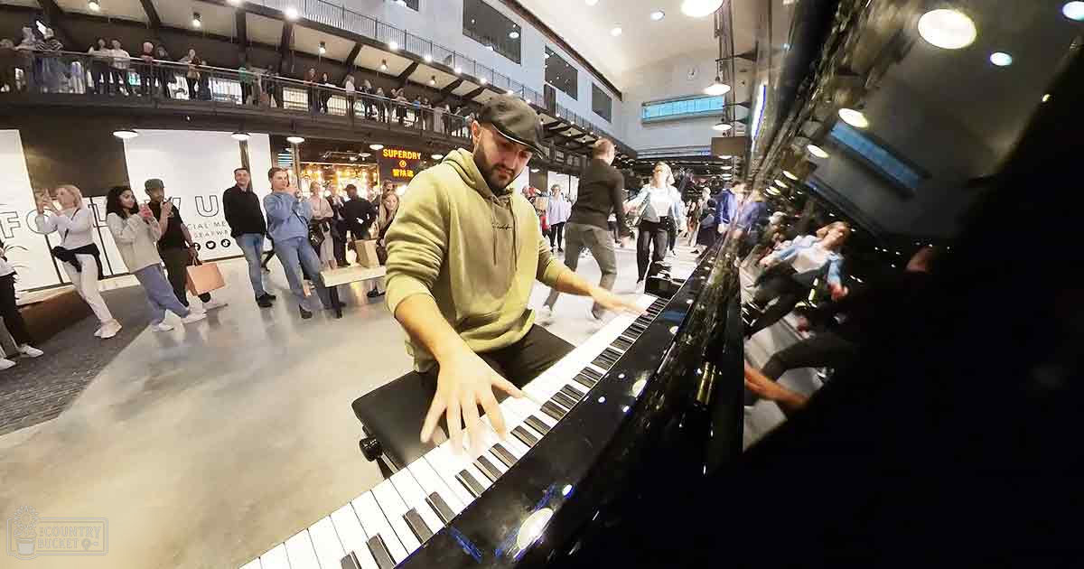 An adult male singer wearing a green hoodie with a hood up, dark pants, and a dark cap plays piano amidst young adults in a multi-level public indoor space with stores below.