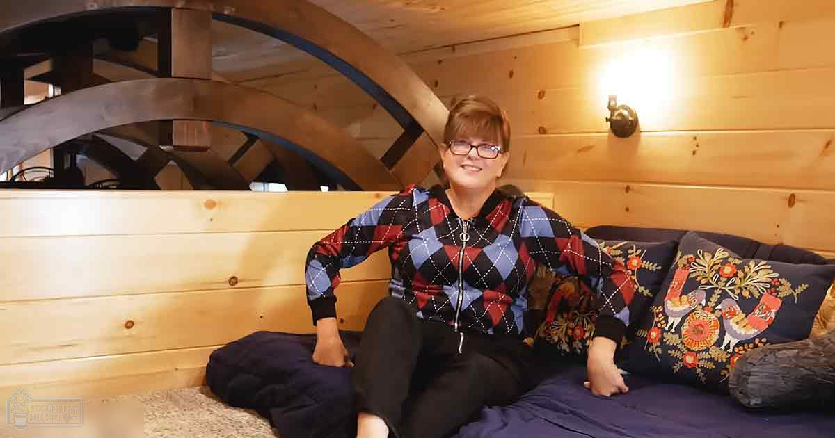 An older adult female in a cozy cabin setting, wearing a zip-up hoodie with a plaid pattern, glasses, and floral pillows inside the rustic interior.