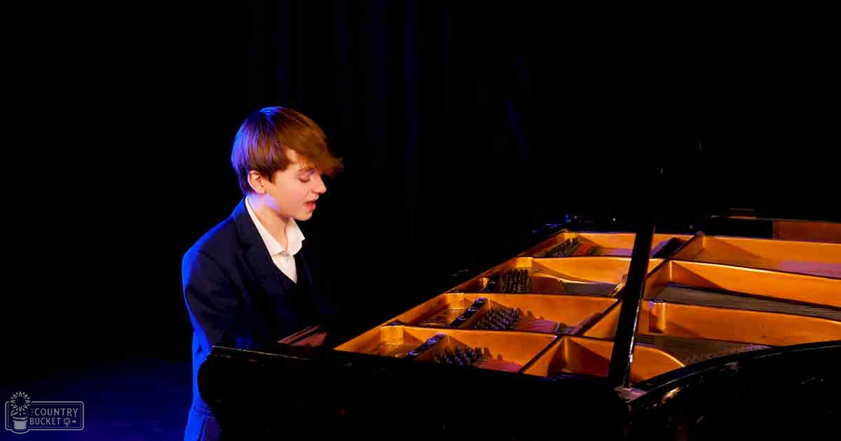 A young adult male in a dark suit jacket plays a grand piano with warm-toned wood, set against dark curtains in an indoor performance hall or studio during what appears to be evening or night time.