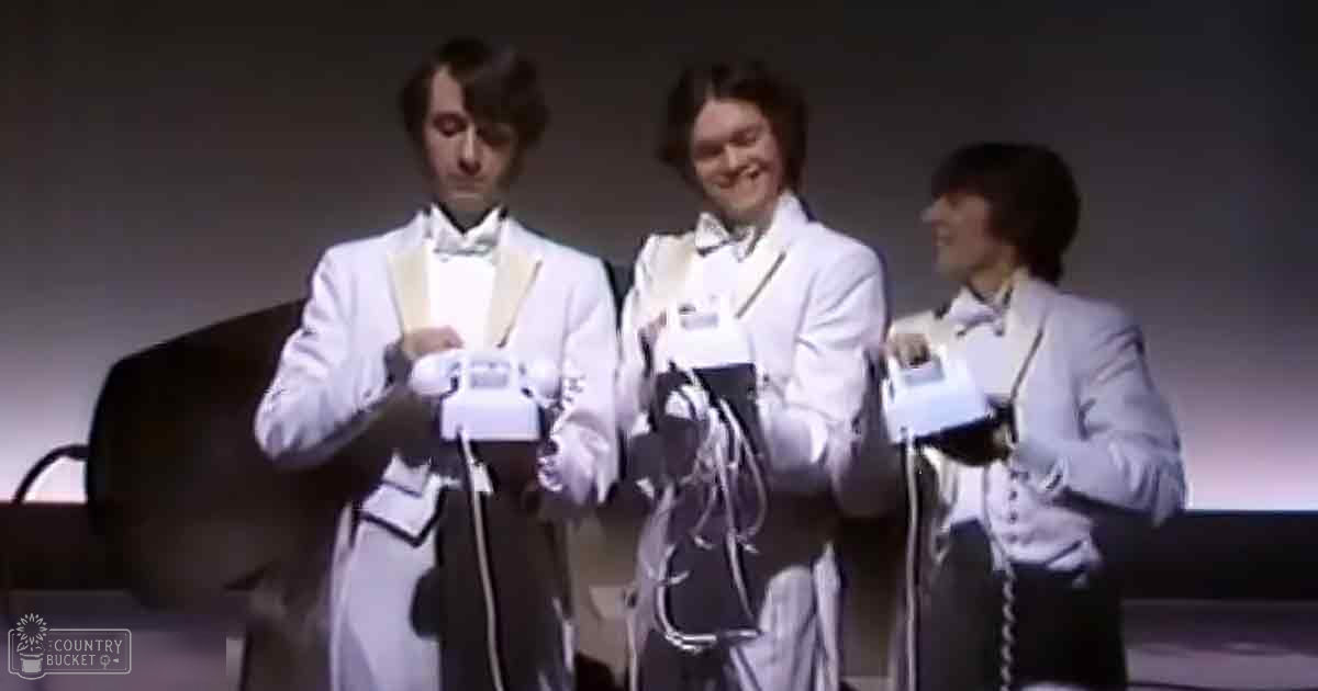 A group of young adult males dressed in formal attire at an indoor event or performance setting; each wearing a white tuxedo, light-colored suit jacket, and holding a device with wires attached.
