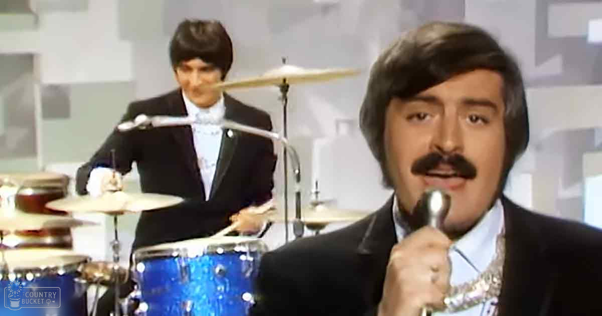 An adult male singer in a dark 1970s-style suit jacket and patterned tie, performing into a microphone with another young adult male drummer in the background wearing a similar vintage outfit.