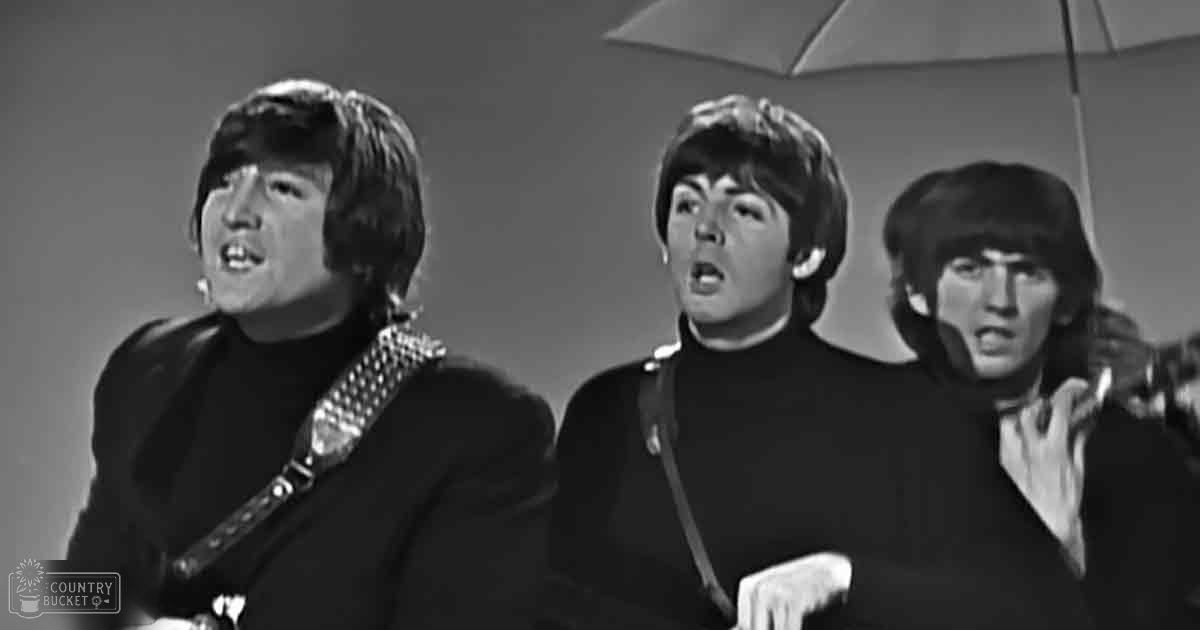 A musical performance scene with a group of young adult males, dressed in black turtleneck sweaters and jackets, set against a simple studio backdrop with neutral-colored walls.