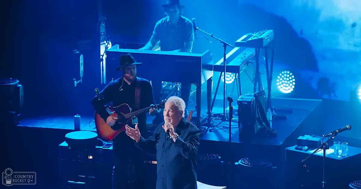 An older adult male singer wearing a dark, buttoned shirt performs on stage with other musicians, including a guitarist and keyboard player in a concert setting under blue stage lights.