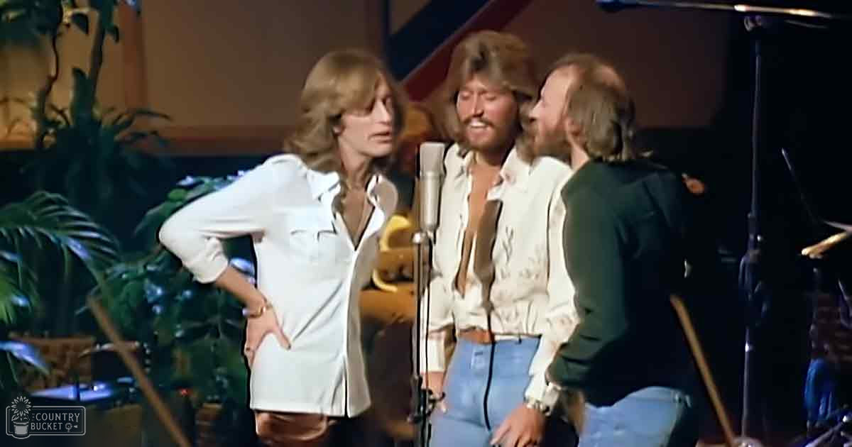 A musical performance scene with three adult males in a studio or stage setting; they are wearing casual and semi-formal attire with focus on white button-up shirts, dark pants, and jeans. The background includes greenery and part of a drum set indicating the setting is for performances.