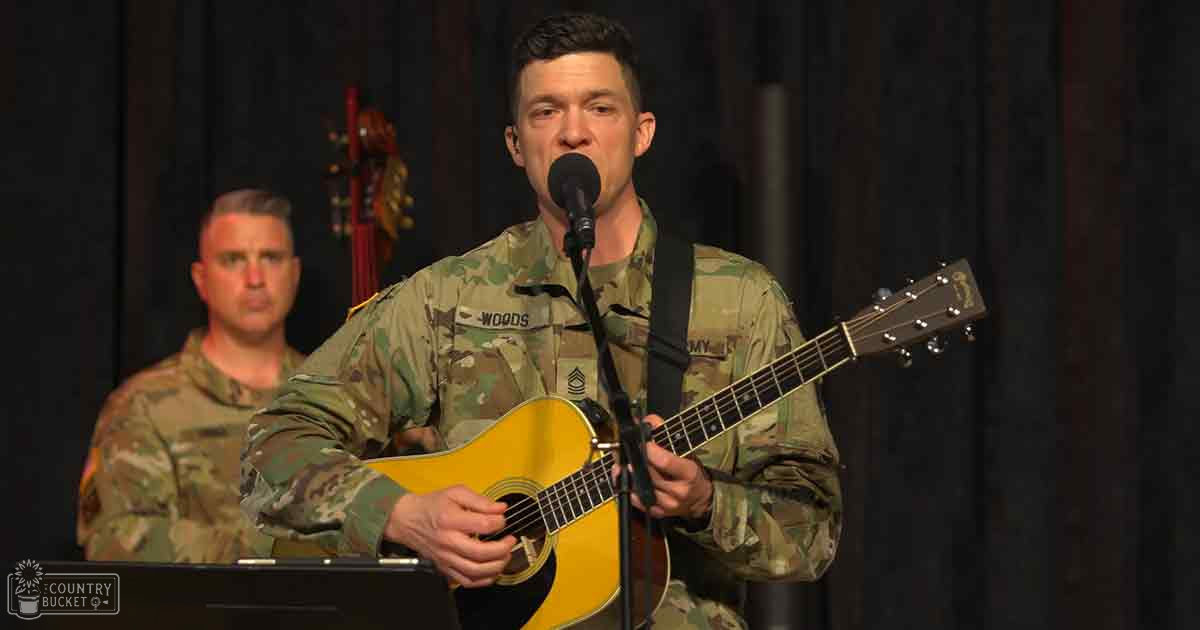 A soldier in a U.S. Army ACU uniform performs an acoustic guitar solo on stage under controlled indoor lighting.