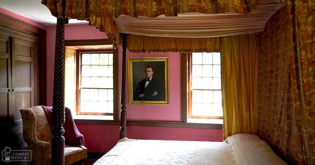 An older-style bedroom with ornate four-poster bed, patterned upholstered chair, ruffled curtains, framed portrait above a bed in natural light.
