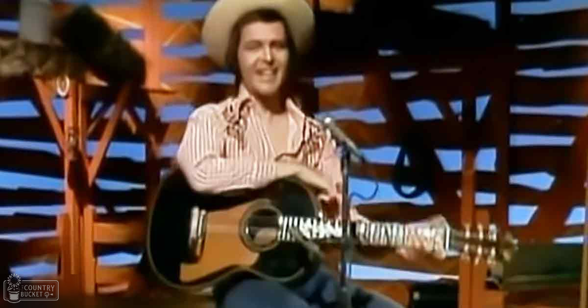 An adult male singer wearing a cowboy hat, striped shirt with fringed details in an indoor stage or studio setting with blue and orange geometric patterns on the backdrop.