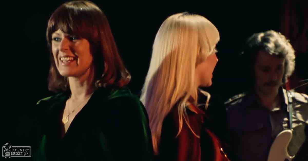 A musical performance scene with three adults, two females and one male, wearing dark green, red jackets, and a black shirt respectively, in an indoor setting with a red curtain and stage-like environment. The scene suggests a performance or recording session.
