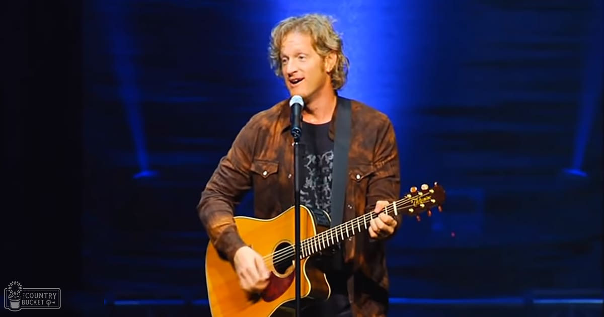 An adult male singer performing on a stage with blue lighting, wearing a brown leather jacket over a black t-shirt with 'Madly Odd' logo in the corner.