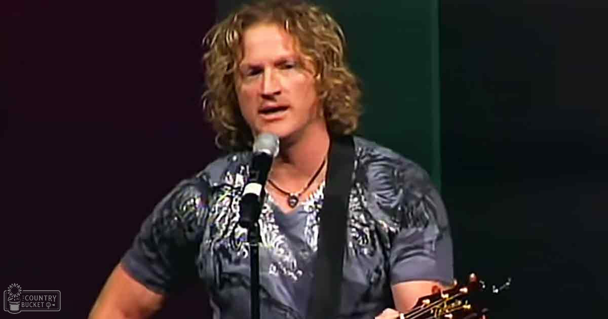 A middle-aged male singer wearing a dark blue shirt with white and light blue patterns on stage, holding a guitar or similar stringed instrument under dim indoor lighting.