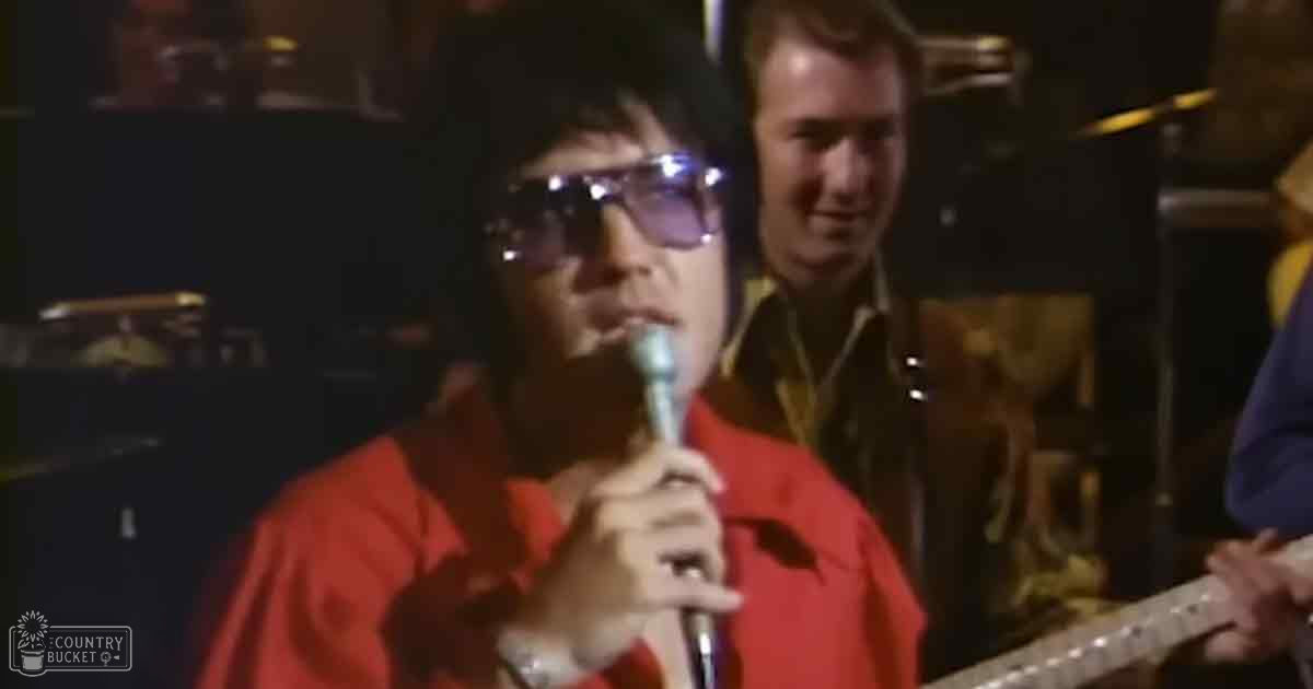 An adult male singer performing into a microphone, wearing a red 1970s-style shirt with a wide collar and long sleeves, surrounded by other performers in similar attire on a dimly lit stage.