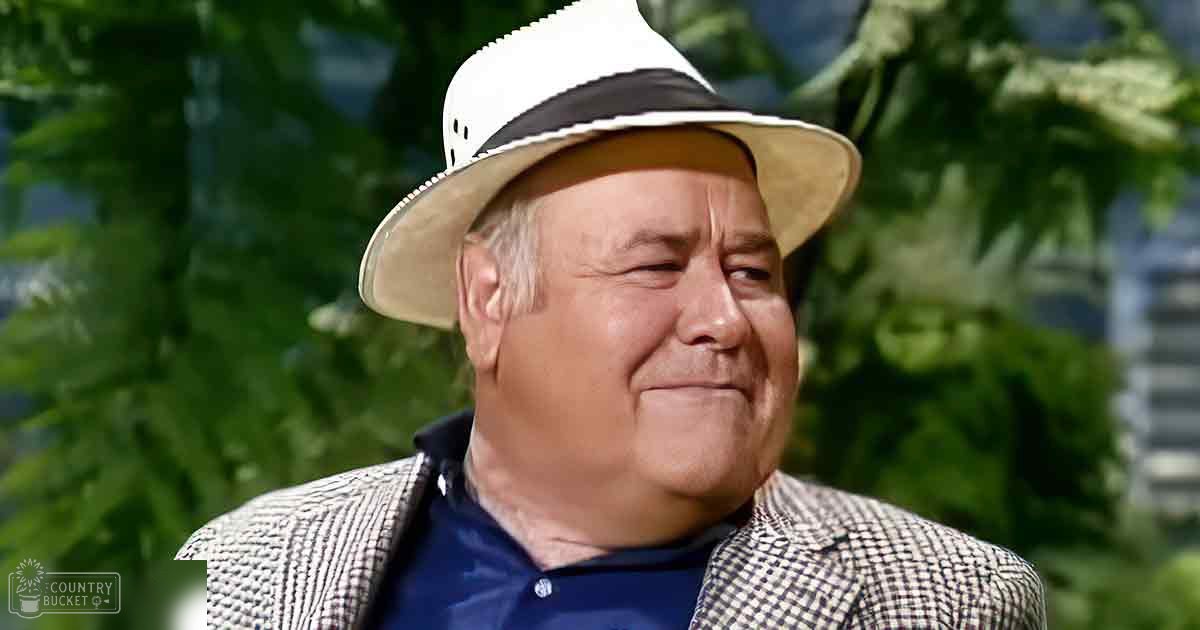 An older adult male in a park-like setting, wearing a white and black checkered blazer over a dark blue collared shirt with a small collar. The scene is outdoors with greenery and trees in the background.