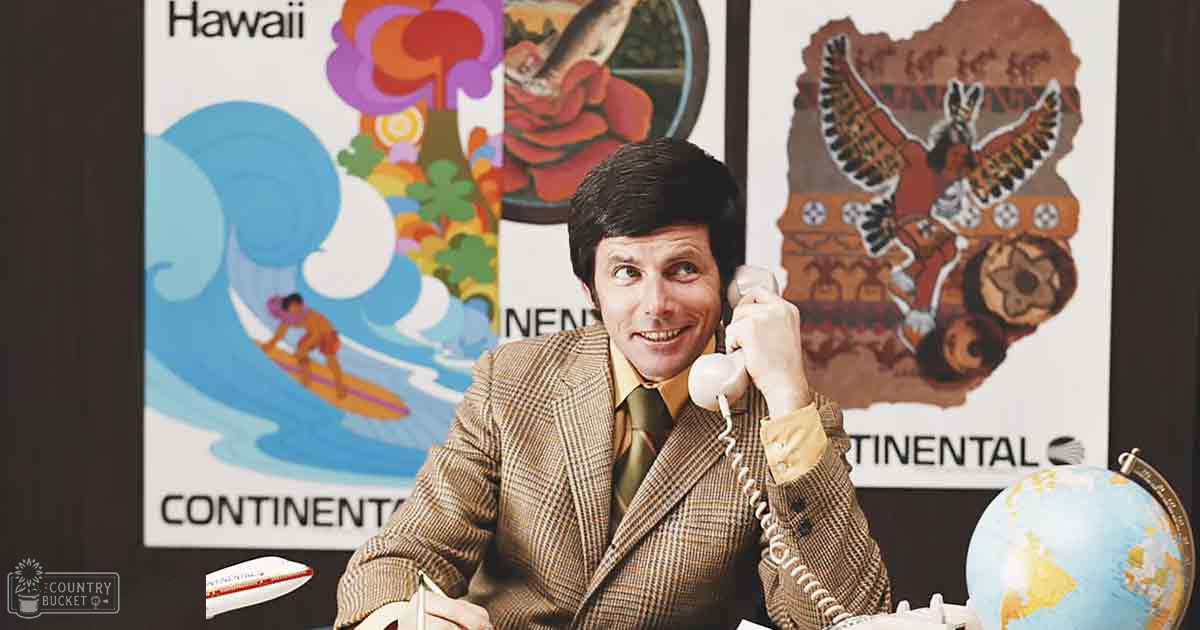 An adult male wearing a tailored brown plaid blazer with a notched lapel, standing in an office setting focused on travel or aviation.