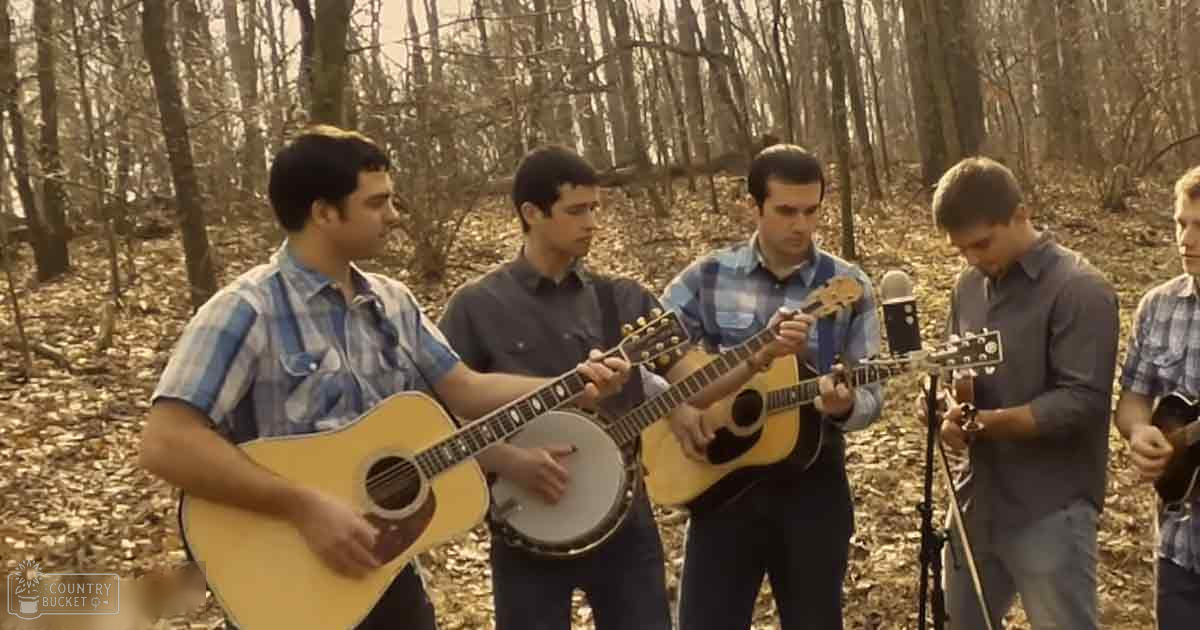 Young adult males playing music outdoors in a forested area with bare trees and fallen leaves; light suggests daytime during autumn or early spring.