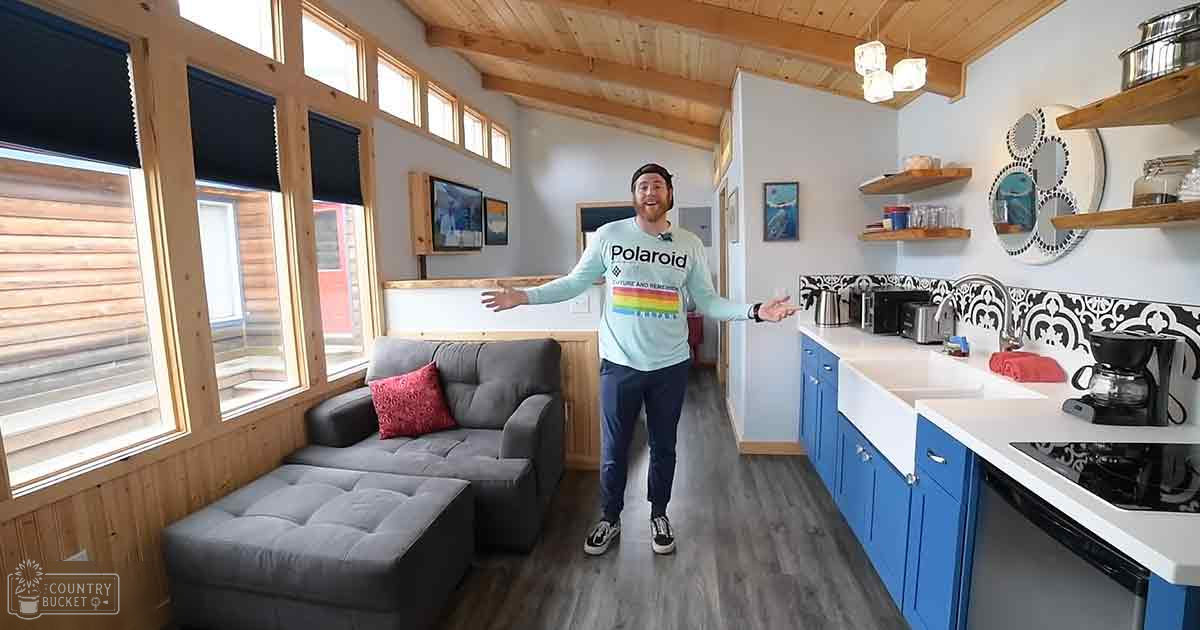 A young adult male wearing a light blue long-sleeved sweatshirt with a rainbow stripe on the chest, paired with black sneakers and a wristwatch, in an indoor cabin setting.