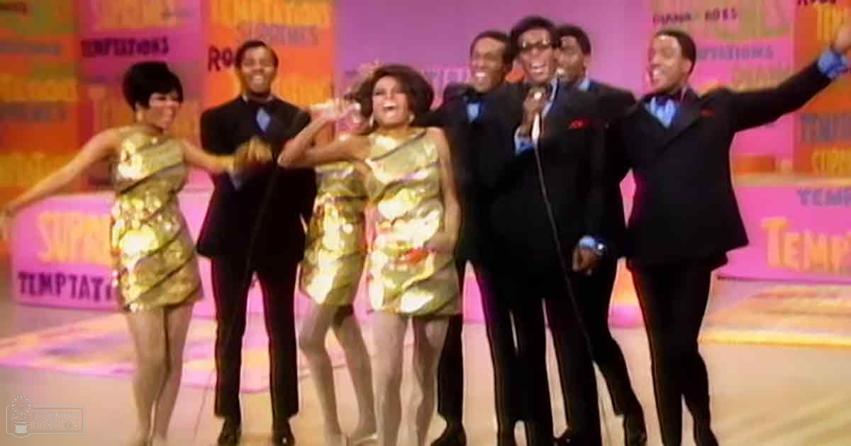 A lively musical performance group from the 1960s or 1970s on a brightly lit indoor stage with colorful banners and text indicating 'Temptations' and 'Rockin'.