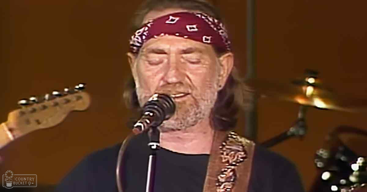 A middle-aged male singer wearing a black t-shirt and red bandana performs on stage, holding a guitar strap over one shoulder. Microphone stand and drum set are visible in the foreground of an indoor music venue with warm lighting.