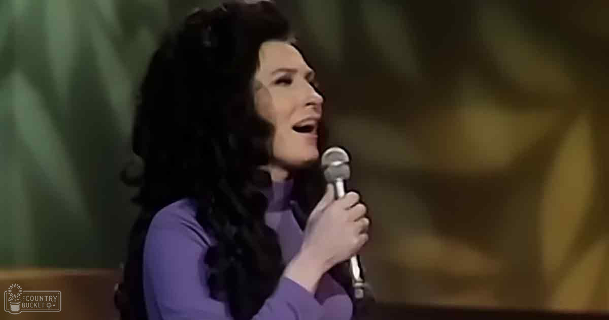 A woman sings into a microphone in what appears to be an indoor setting, wearing a long-sleeved purple top with a high neckline and fitted sleeves.