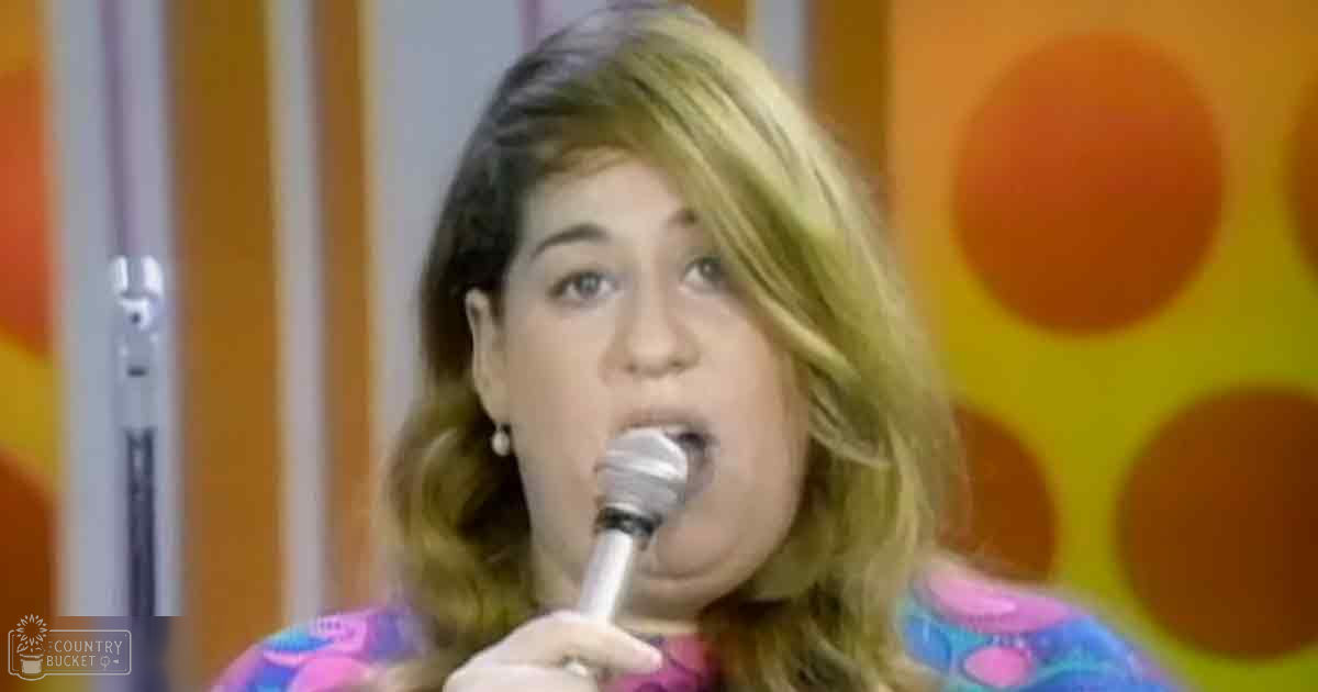 A middle-aged adult female singer wearing a multicolored patterned long-sleeved top, holding a microphone on stage with vibrant orange and yellow circular patterns in the background.