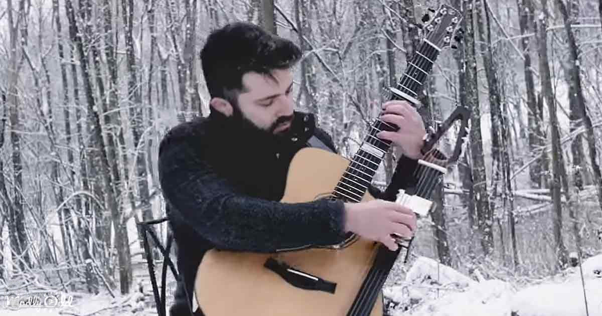 Adult male playing an acoustic guitar amidst a snowy forest setting with bare trees covered in snow.
