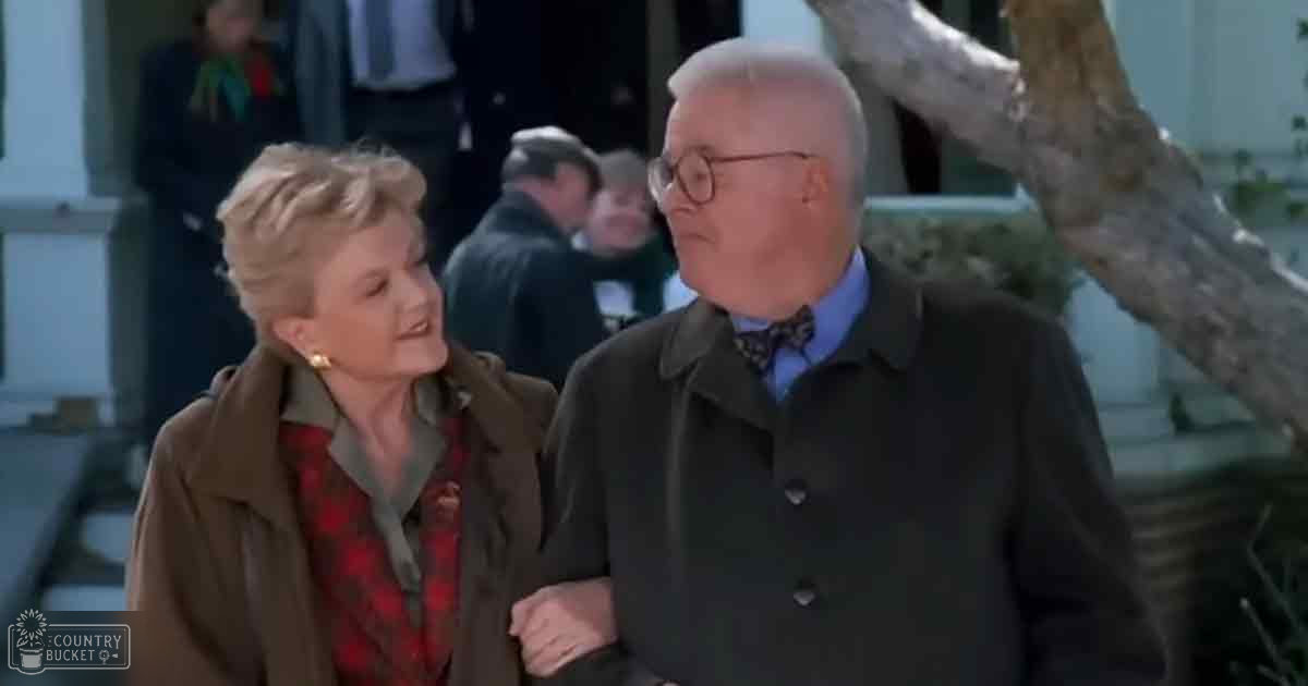 An older adult couple wearing mid-20th century attire outdoors, surrounded by a public setting with buildings and trees.