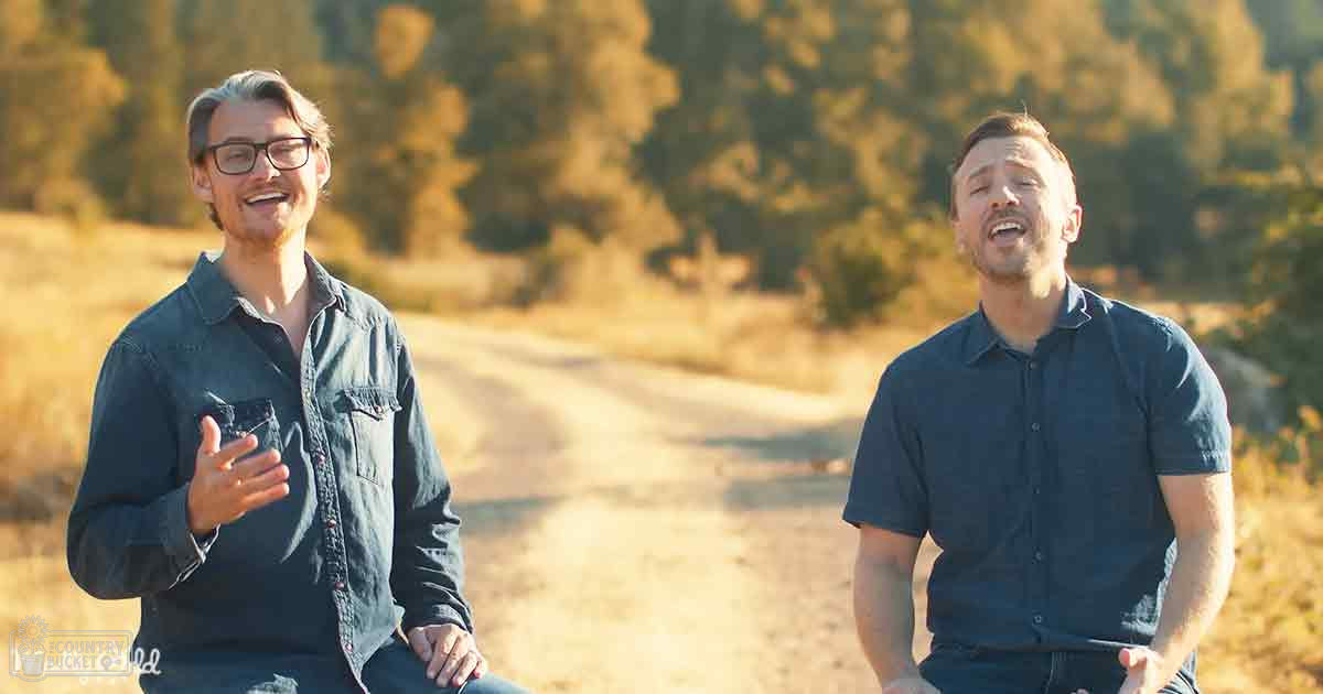 Two young adult males smile outdoors on a path surrounded by trees with warm sunlight filtering through the leaves.