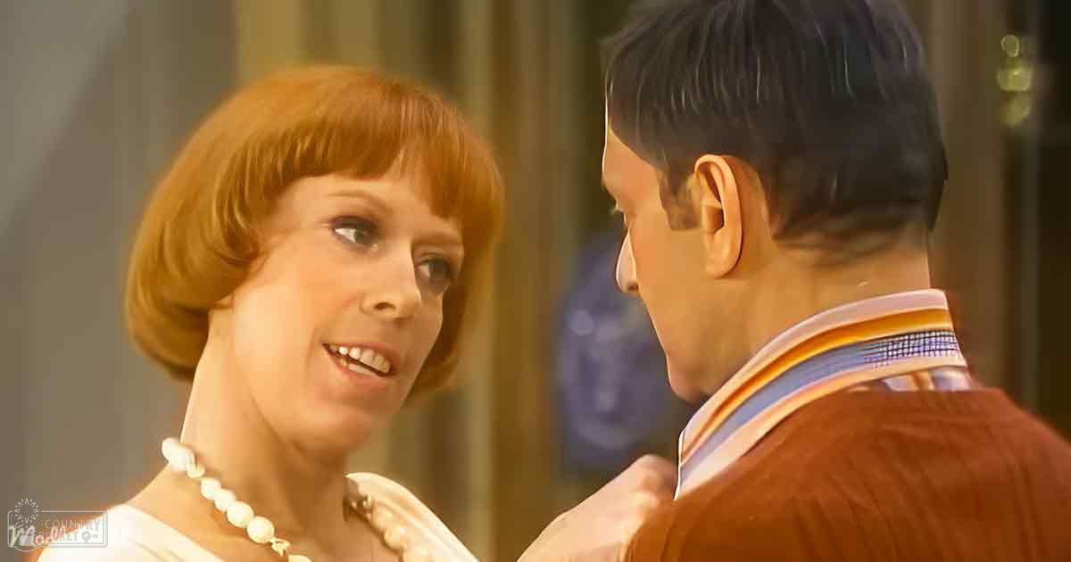 A woman wearing a pearl necklace and a white garment with high collar stands indoors near a man in an orange sweater and striped shirt.