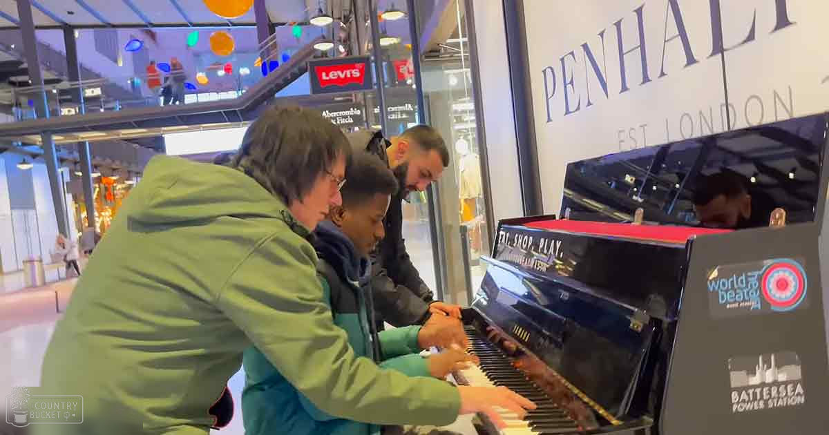 A group of three adults playing a piano in an indoor shopping mall with bright artificial lighting.