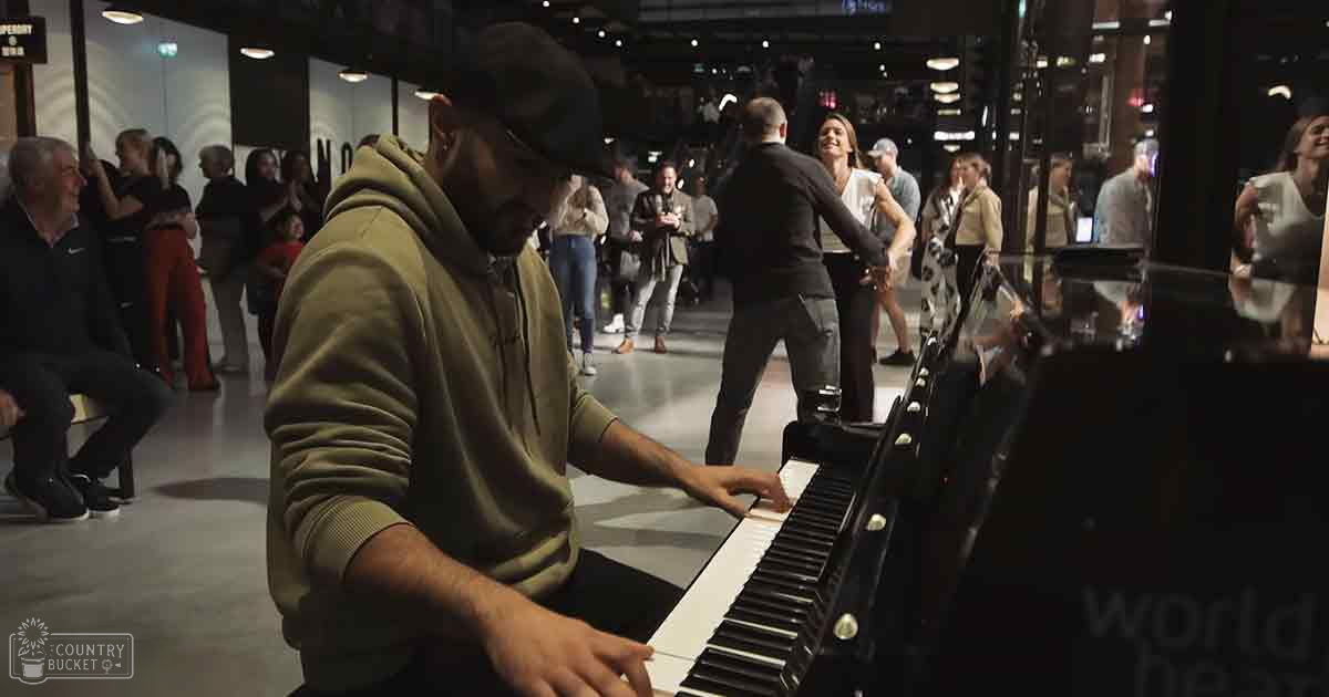 Adult male playing piano in modern indoor space with glass walls and metal structures, surrounded by other people engaged in various activities under artificial lighting.