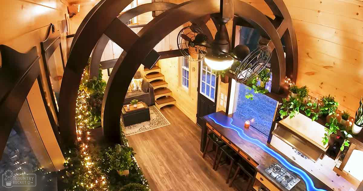 A cozy cabin interior with a spiral staircase, decorative fan, Christmas tree adorned with lights and ornaments, kitchen area, potted plants, warm lighting during evening or nighttime.