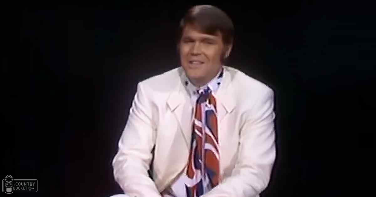 A middle-aged adult male in a formal white suit with a patterned tie stands against a dark backdrop, possibly on stage.