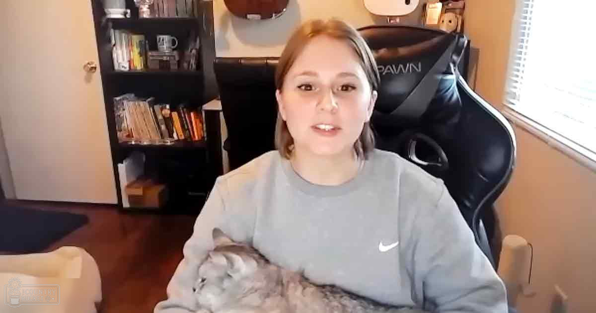 A young adult female wearing a gray Nike sweatshirt sits with a tabby cat on her lap in an indoor room filled with bookshelves and natural light.