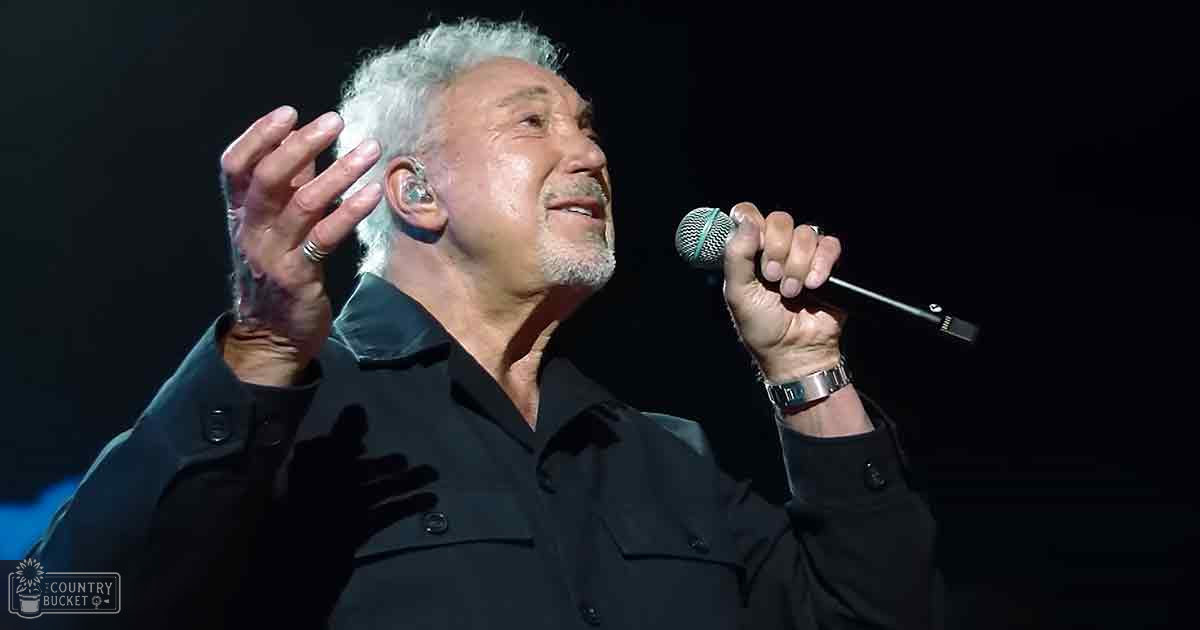 A middle-aged man in a black button-up shirt performs on stage, holding a microphone with a silver wristwatch visible. The setting is dimly lit and appears to be an indoor performance venue.