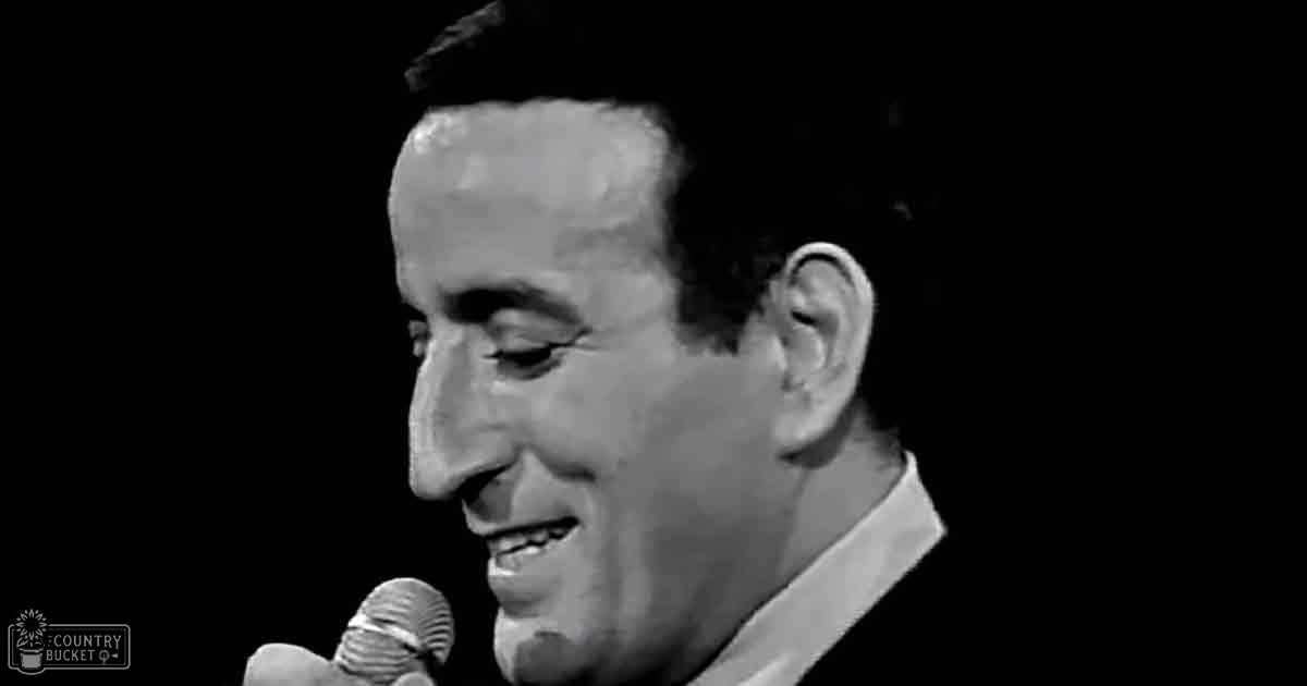A middle-aged male performer wearing a white collared shirt with 'Madly Odd' text, holding a microphone in front of him against a black studio background.