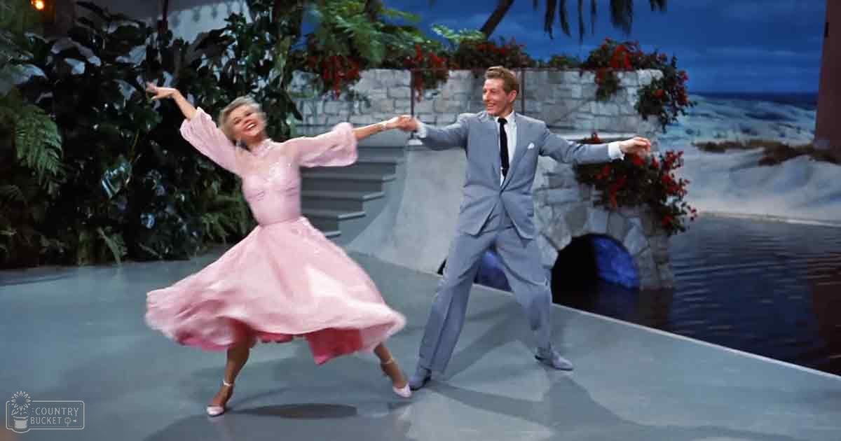 A couple dancing on a stage with lush greenery and beach scenery in the background, suggesting an outdoor setting for a musical or theatrical performance.