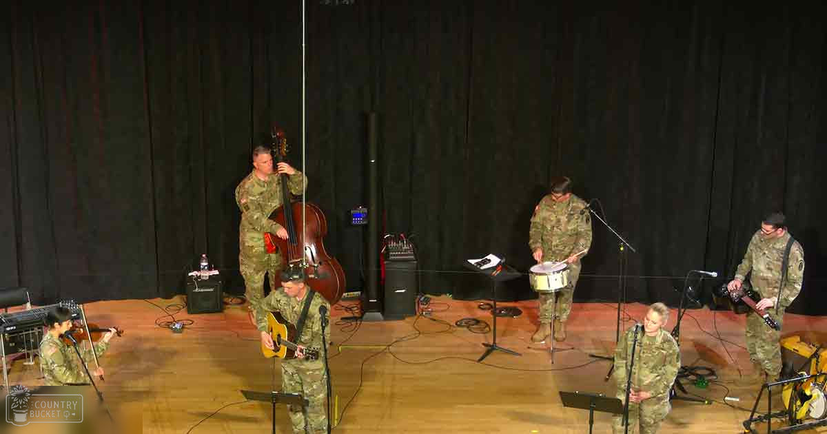 A military band performing indoors with adult males in camouflage uniforms playing instruments like double bass, acoustic guitar, drums, and electric guitar; dark curtains and wooden flooring suggest a theater or auditorium setting.