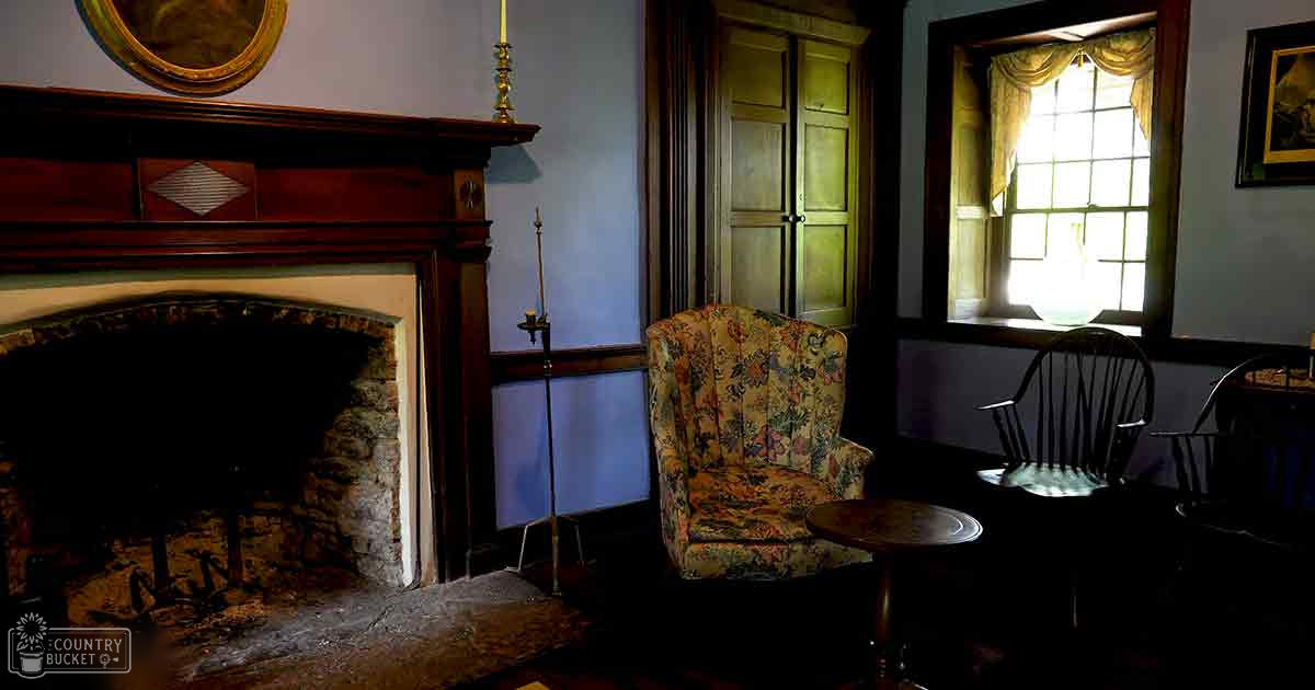 An ornate floral-patterned armchair sits beside a fireplace in an old, historic home with light blue walls and dark trim, wooden double doors, and a window with curtains. The setting is indoors during the daytime.