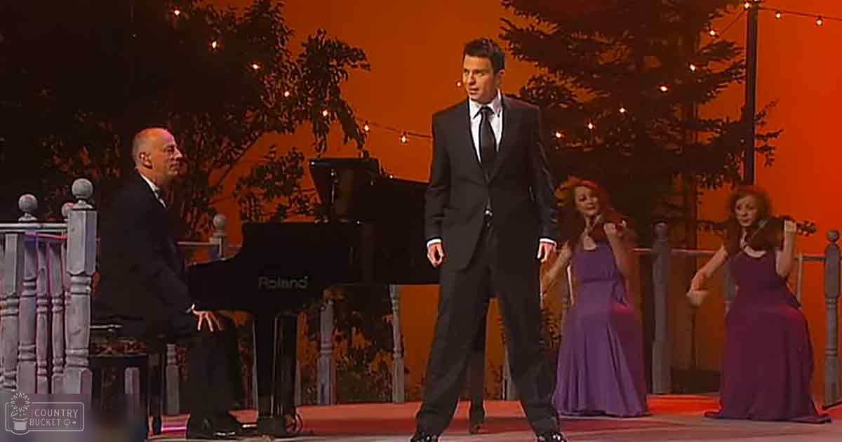 A male adult singer in a dark suit and white shirt performing on stage with two female violinists in purple dresses, accompanied by an older adult pianist at a Roland piano. The setting is an indoor stage performance with orange-toned lighting and tree silhouettes.