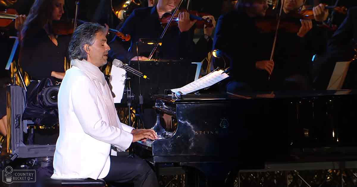 A man playing piano amidst an orchestra in a dimly lit indoor concert hall, surrounded by musicians and stage equipment.