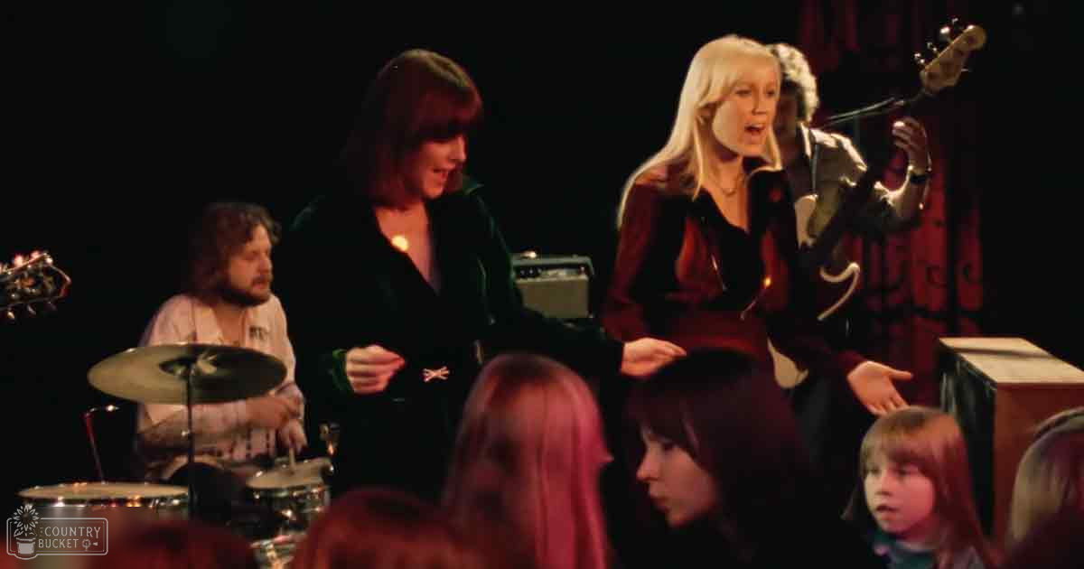 Musical performance by an adult male singer with 'PIT' text shirt playing drums; other performers wearing dark outfits in a dimly lit indoor venue with stage lighting.