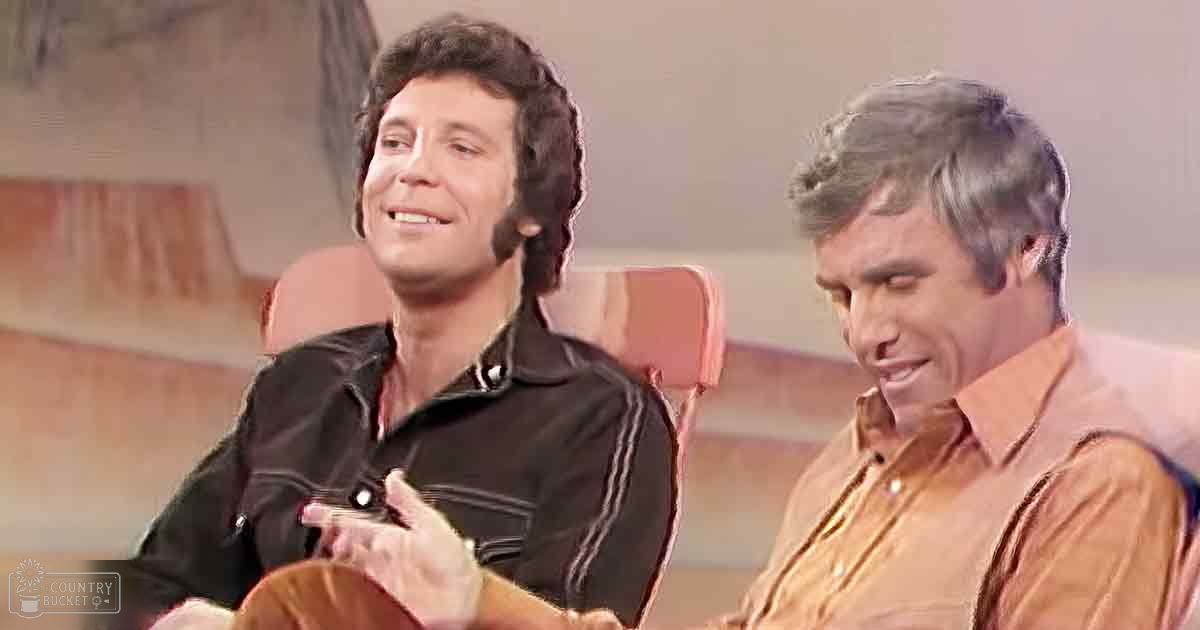 Two adult male individuals seated, one in a black leather jacket with white stripes and the other in an orange button-down shirt with vertical stripes, set against red chairs under bright artificial lighting.