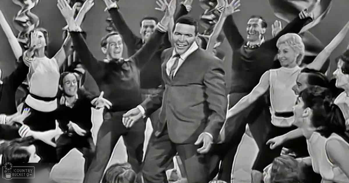 Adult male singer in dark suit with formal 1950s attire, surrounded by young adults in light dresses at a lively indoor performance or celebration.