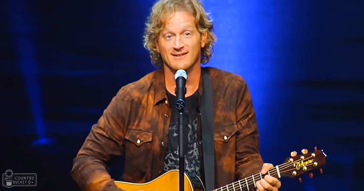A middle-aged male performer in a dark indoor concert venue, wearing a brown leather jacket over a dark shirt with 'Columbia' text and patterns.
